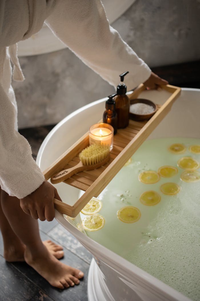 why-choose-me Unrecognizable barefoot female in bathrobe putting wooden tray with spa supplies on tub with lemon slices on water surface during skincare routine